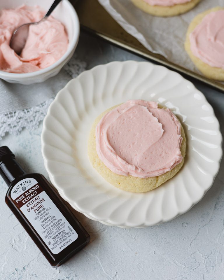 A bottle of almond extract next to a pink-frosted sugar cookie on a stack of small plates.