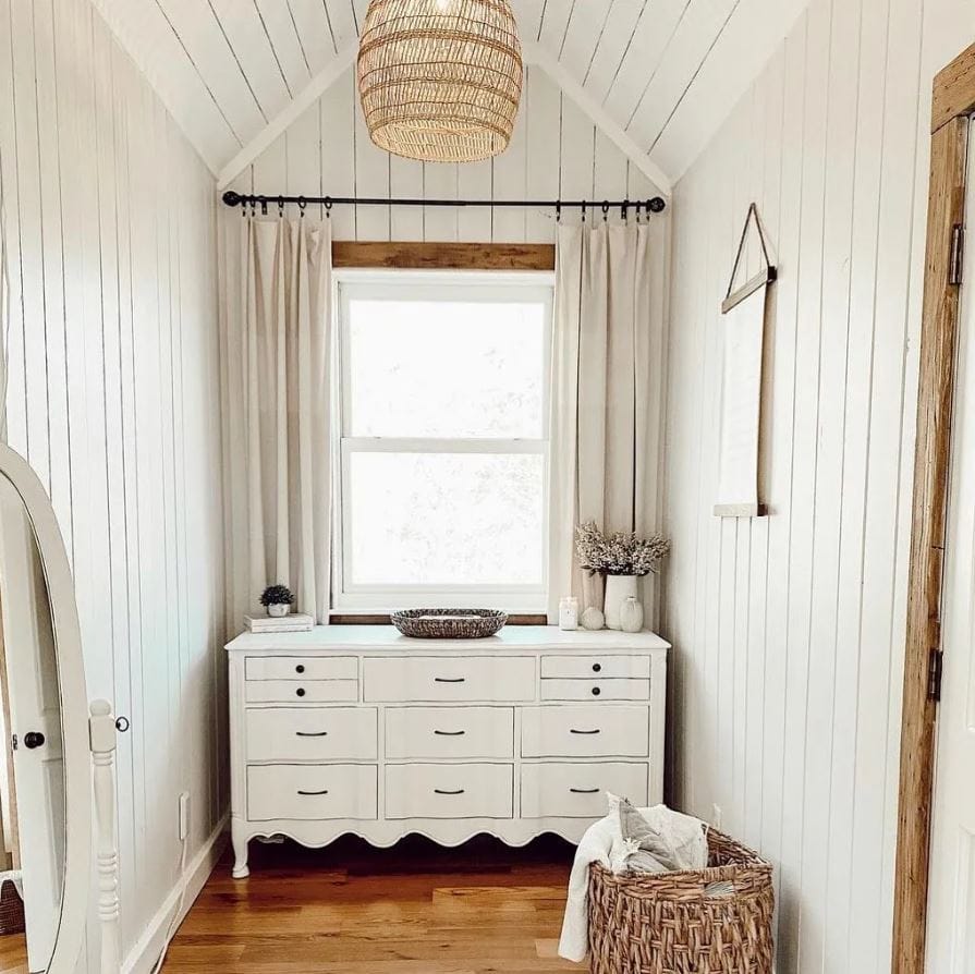 An alcove with vertical white paneling, a planked ceiling, and white sideboard in front of a large window.