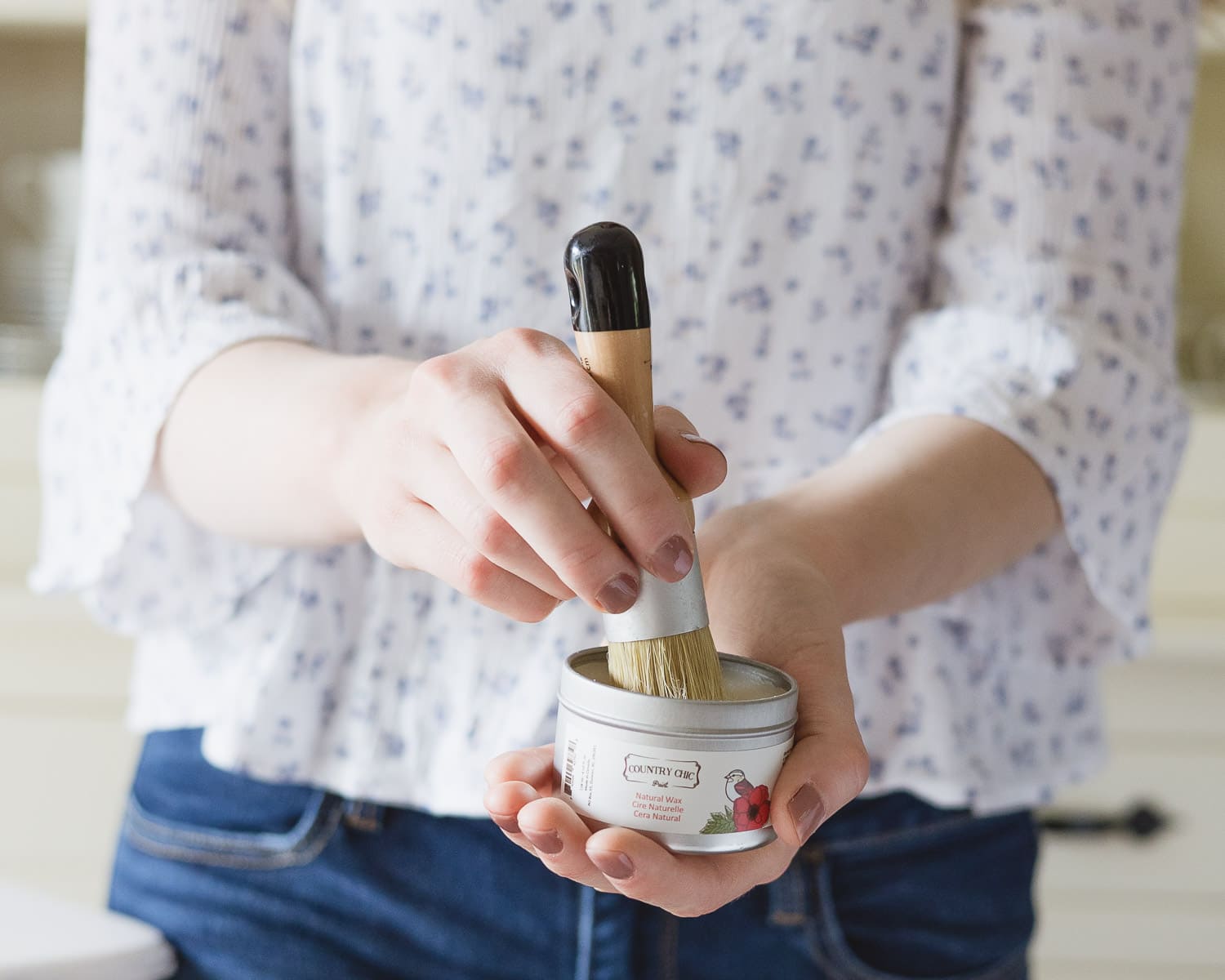 Woman holding a small container of furniture wax and swirling a circular brush inside the container.