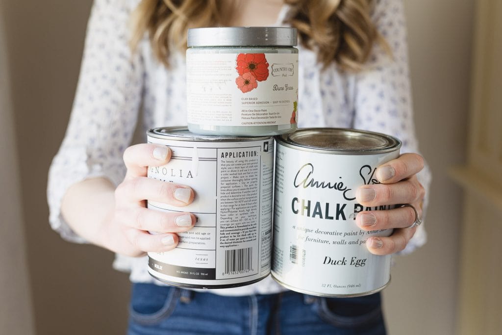 Woman holding three cans of Chalk Paint and chalk style paint from different brands.