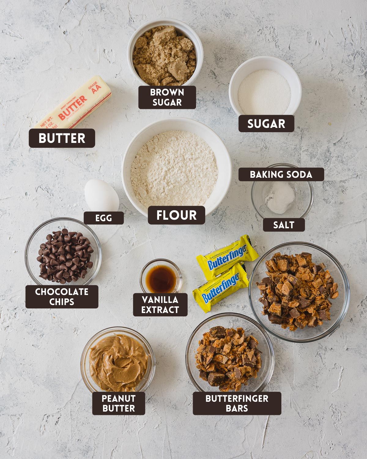 Labelled ingredients for Butterfinger cookies: butter, brown sugar, sugar, egg, flour, baking soda, salt, chocolate chips, vanilla extract, peanut butter, chopped Butterfinger bars.