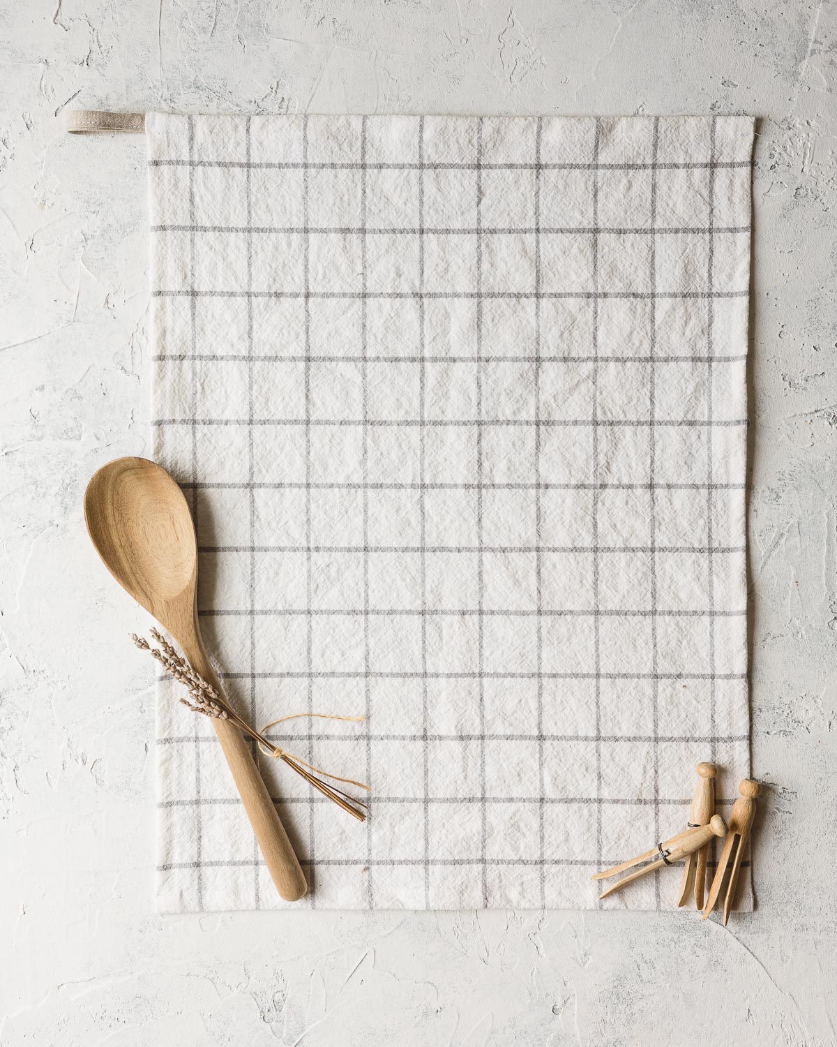 A handmade kitchen towel of linen with a gray grid design and a wooden spoon, lavender, and vintage clothespins displayed on top.