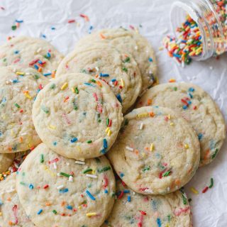 A pile of funfetti cookies on a piece of parchment paper with a jar of spilled sprinkles in the background.