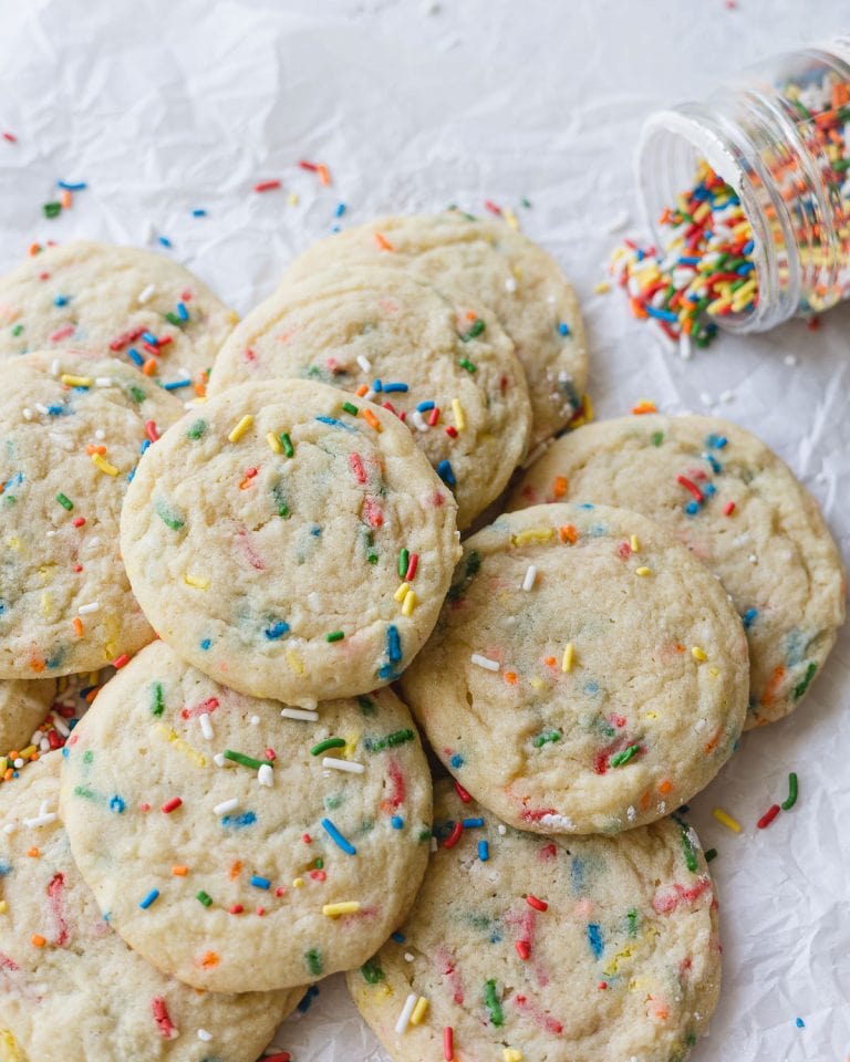 A pile of funfetti cookies on a piece of parchment paper with a jar of spilled sprinkles in the background.
