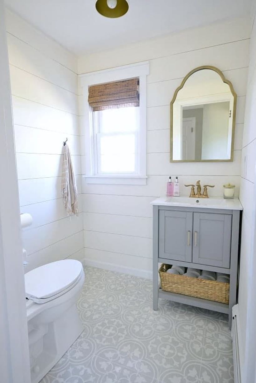 A powder room with shiplap planked walls, a small gray vanity, and a window with farmhouse trim.