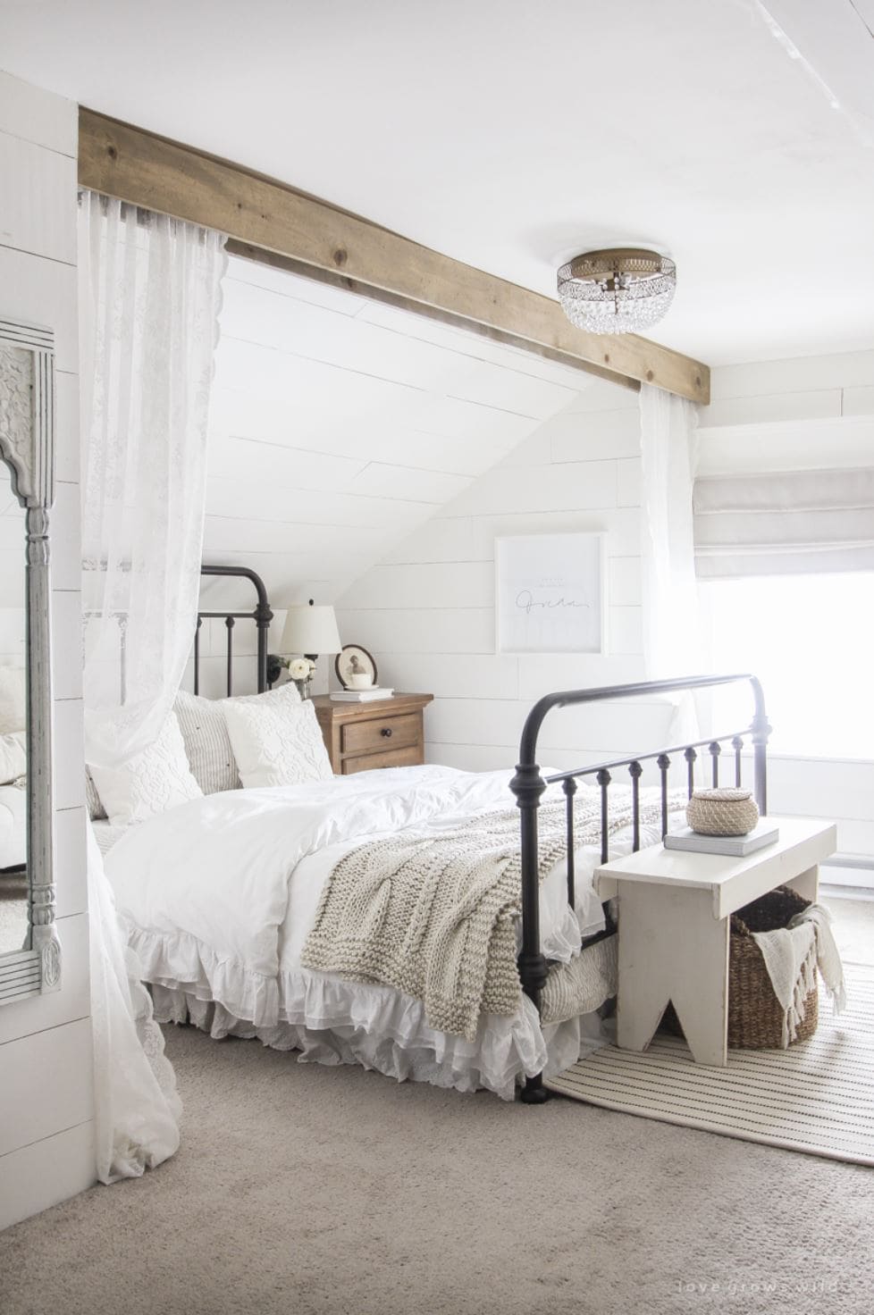 A feminine bedroom with white shiplap walls, lacy bedding and a beam with lace curtains hanging down.