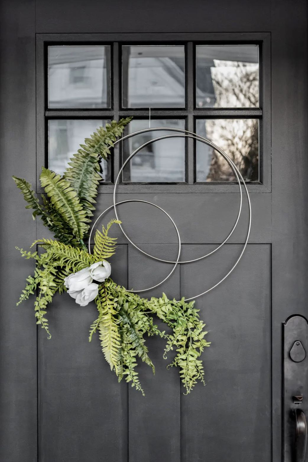 A modern wreath with three hoops, greenery, and white flowers hanging on a black front door.