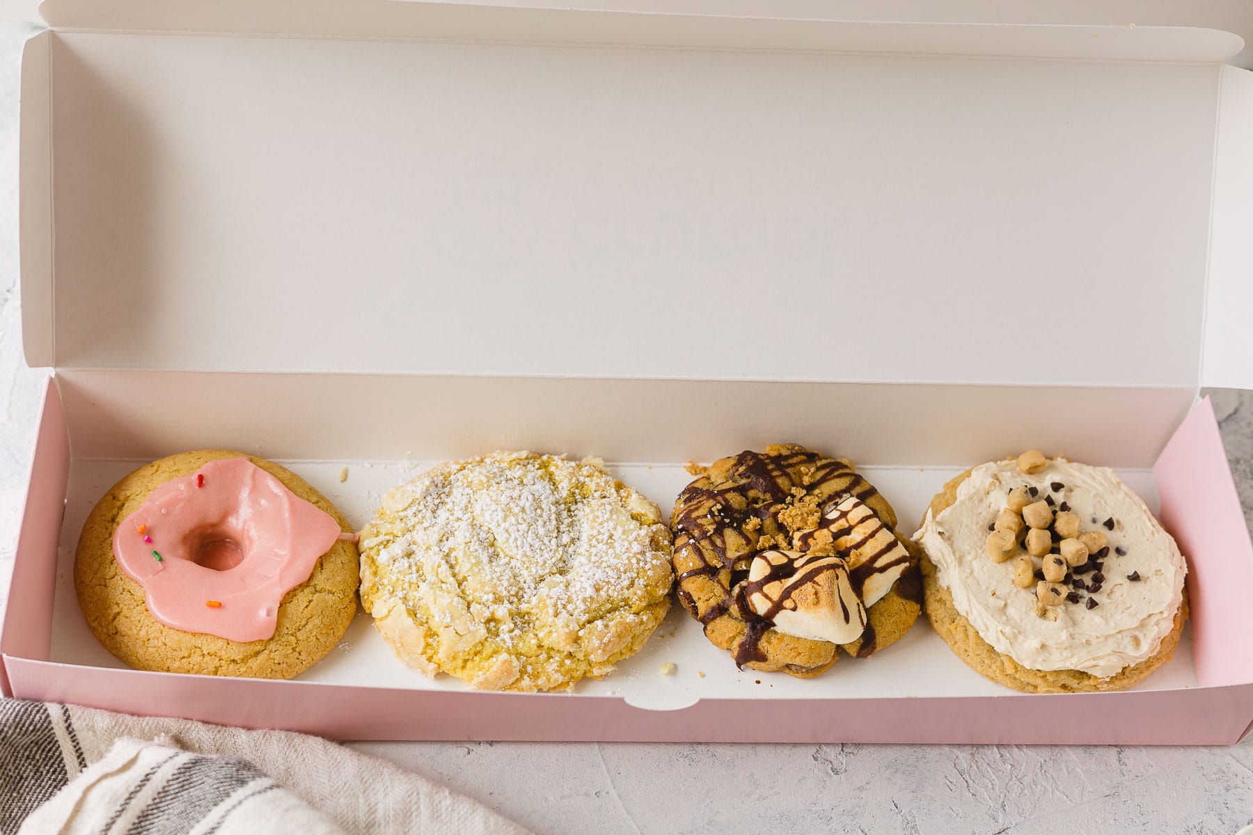 A box of four Crumbl cookies: Pink Doughnut, Lemon Crinkle, S'mores, and Frosted Chocolate Chip Cookie Dough.