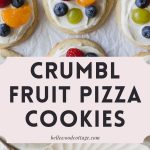 Sugar cookies topped with cream cheese frosting, blueberries, raspberries, green grapes, and mandarin orange slices with the words, "Crumbl Fruit Pizza Cookies."