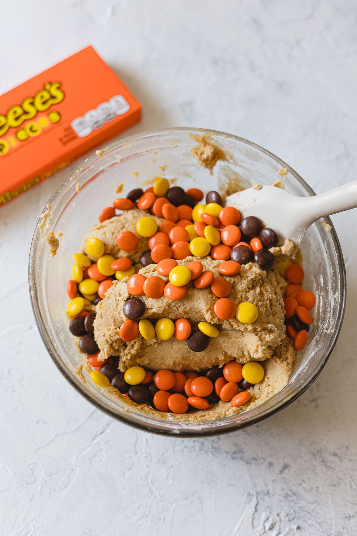Mixing Reese's Pieces into peanut butter cookie dough in a glass bowl.