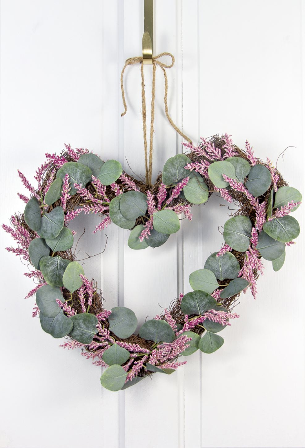 A heart shaped wreath with pink-purple florals and greenery hanging on a front door.