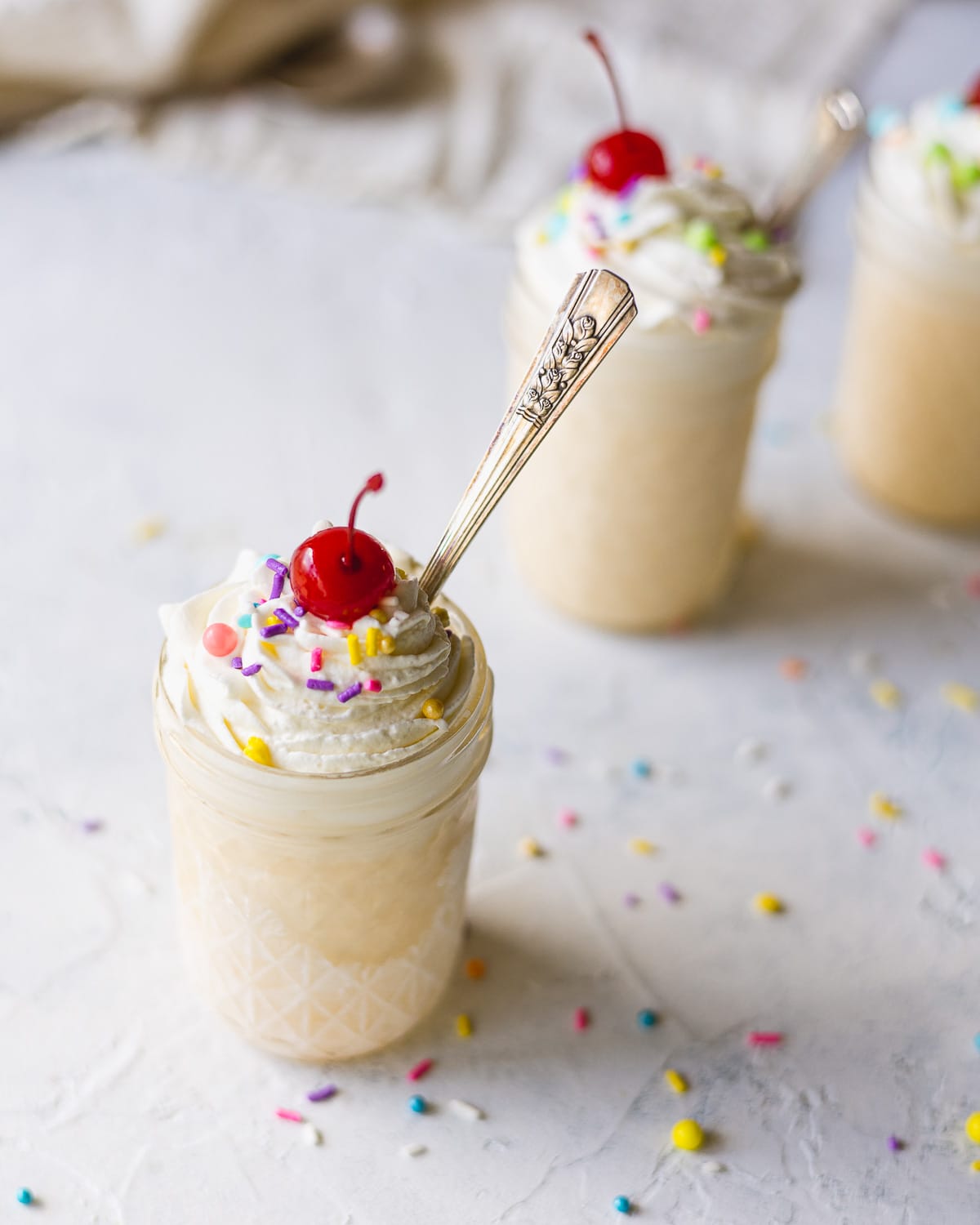 Vanilla milkshake with a swirl of whipped cream, sprinkles, cherry, and spoon.