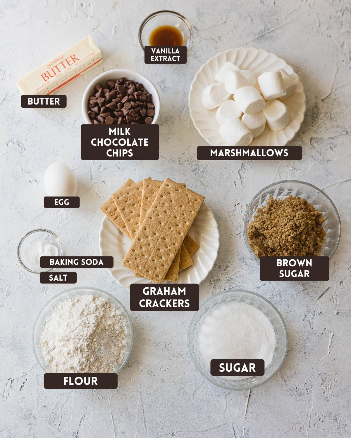 Labeled ingredients for s'mores cookies: butter, vanilla extract, milk chocolate chips, marshmallows, egg, baking soda, salt, graham crackers, brown sugar, flour, and sugar.
