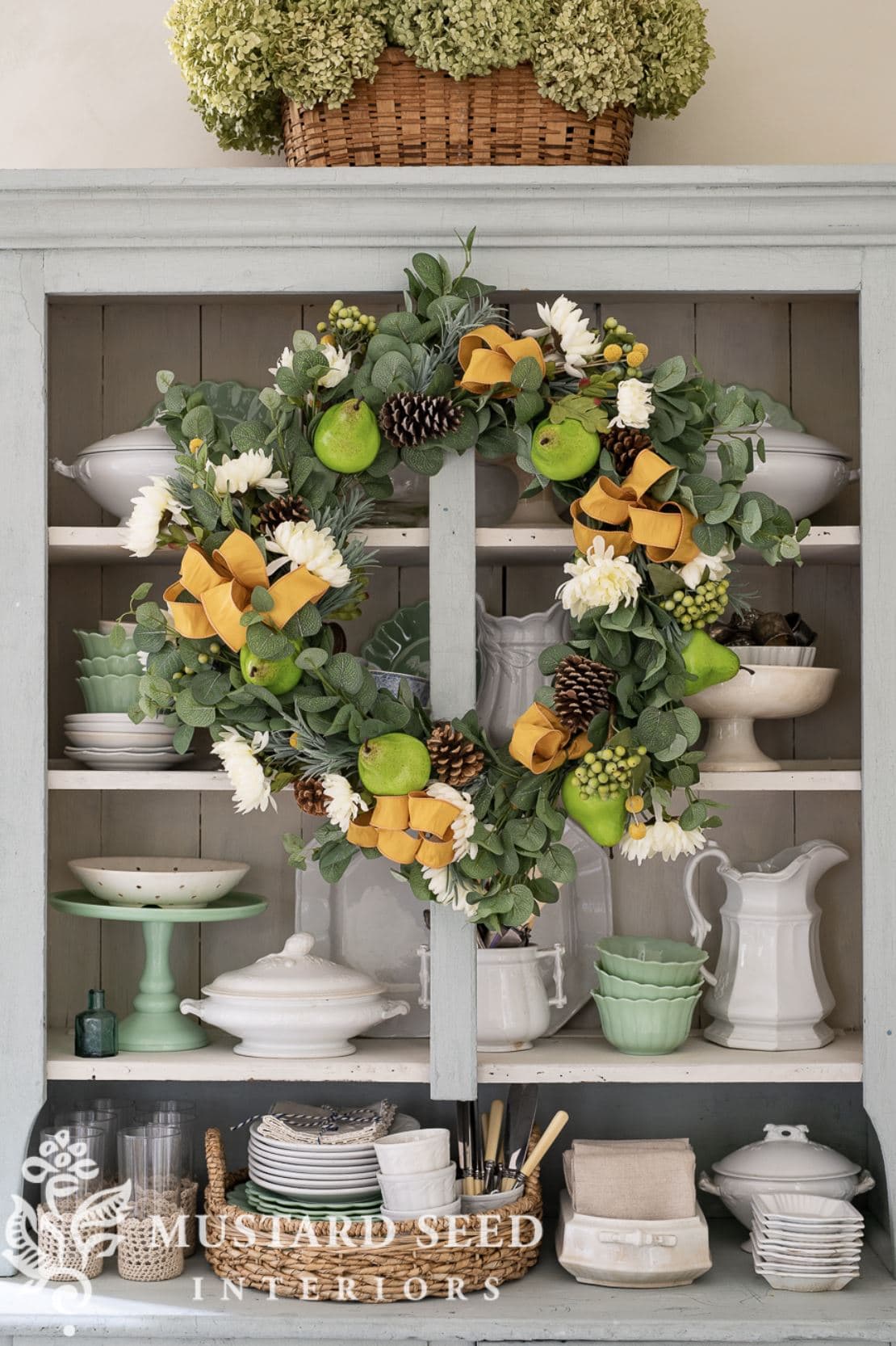 A fall wreath with greenery, orange ribbon, and faux pears hanging over an antique hutch filled with ironstone.