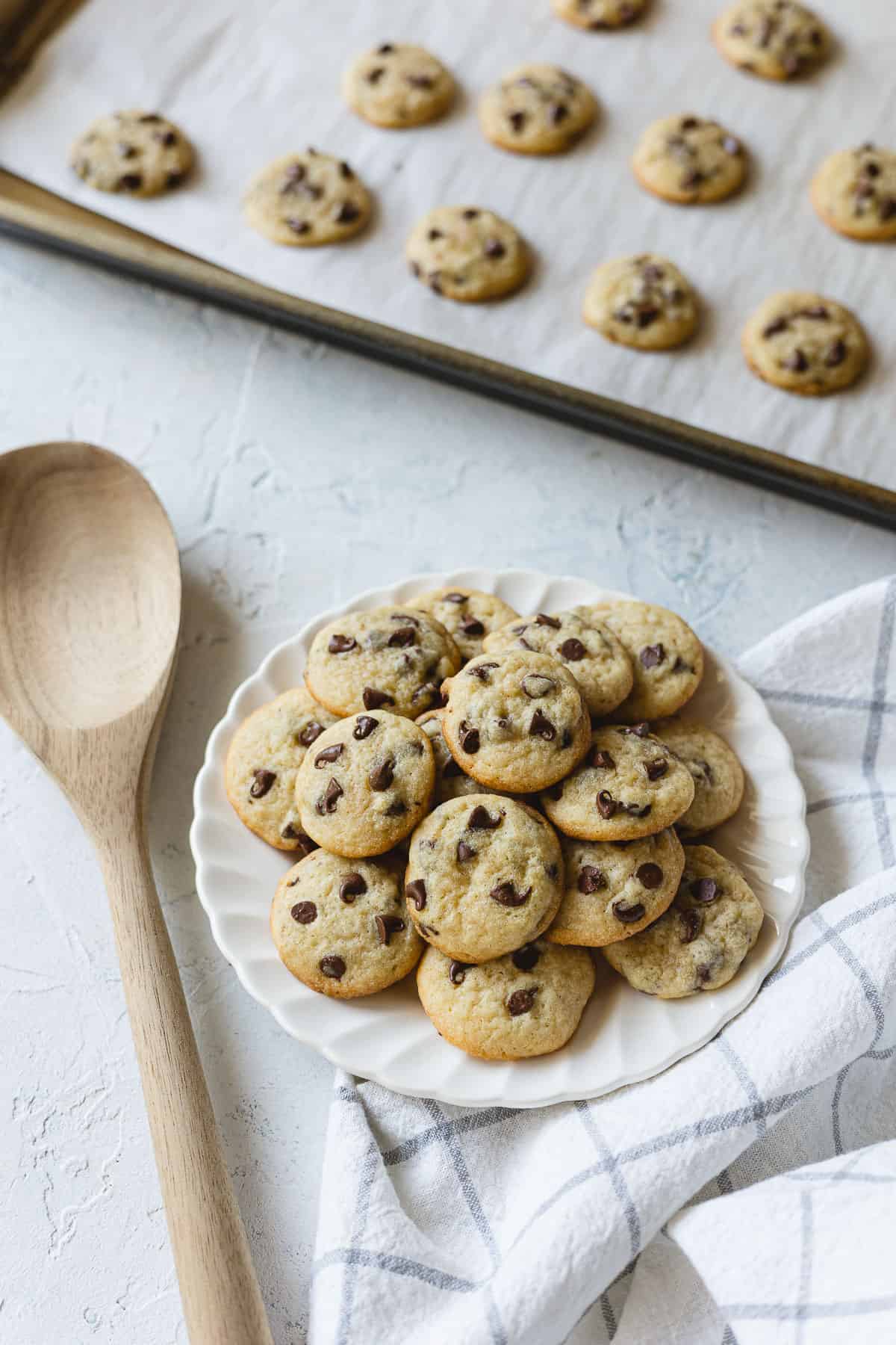 Mini chocolate chip cookies piled on a plate with a wooden spoon and blue-and-white towel.