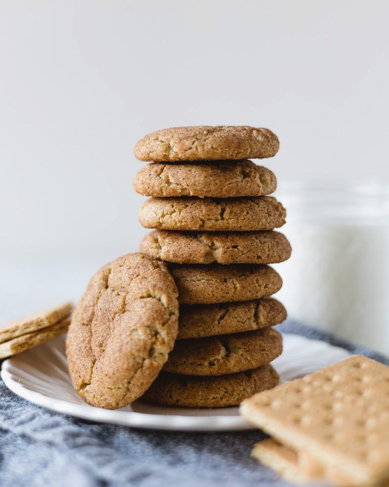 A stack of cookies on a white plate with graham crackers and a glass of milk in the background.