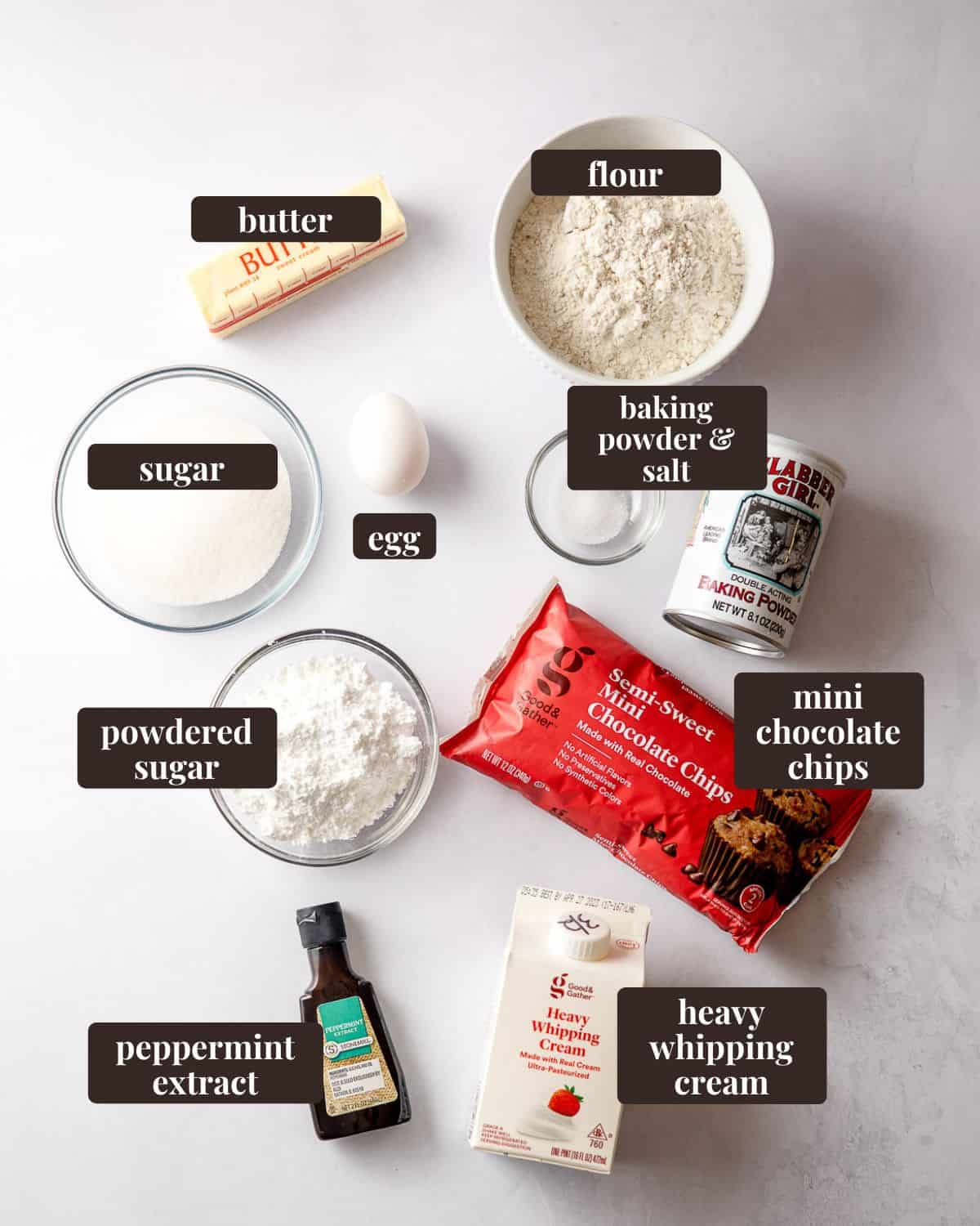 Ingredient arranged and labeled for mint chip cookies: butter, flour, sugar, egg, baking powder and salt, powdered sugar, mini chocolate chips, peppermint extract, and heavy whipping cream.