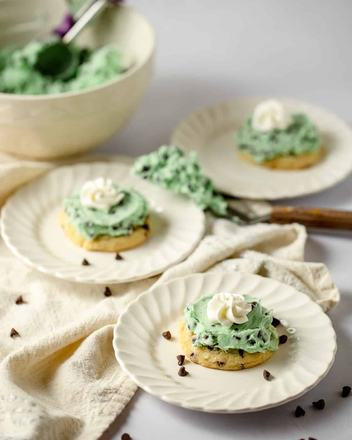 Three plated chocolate chip sugar cookies frosted with green mint buttercream and whipped cream with frosting bowl and spatula in the background.