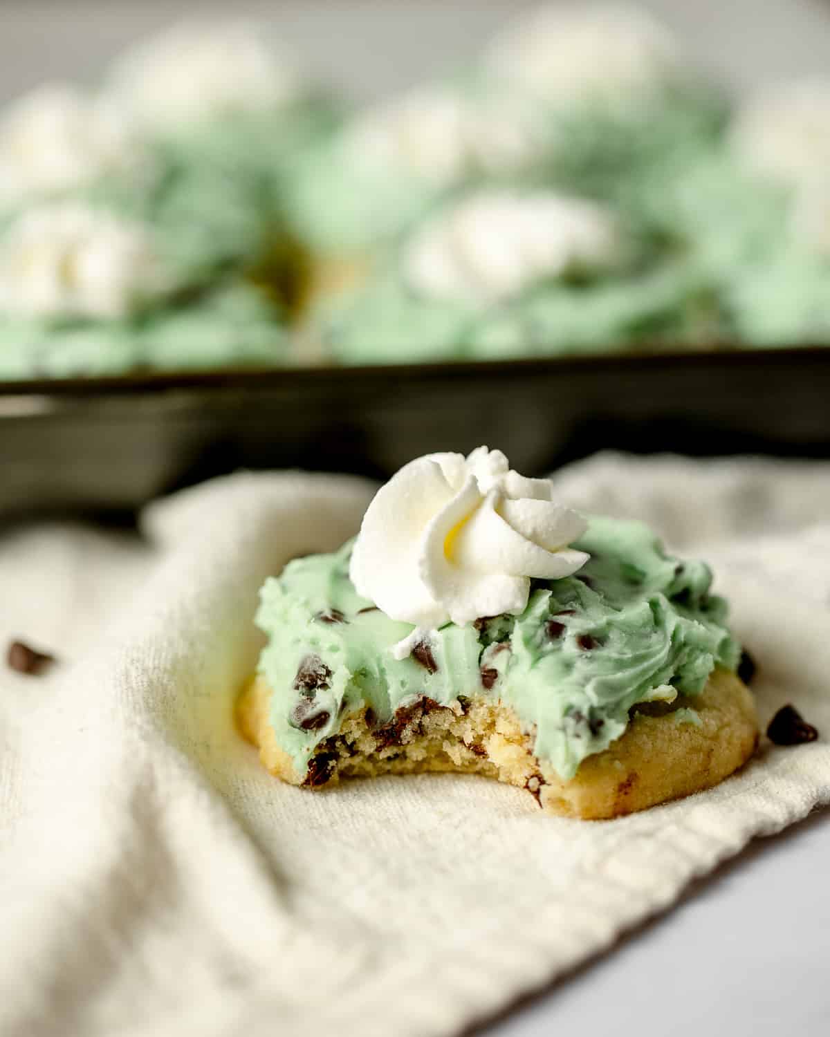 A chocolate chip sugar cookie frosted with mint green chocolate chip frosting and a rosette of whipped cream with a bite removed.
