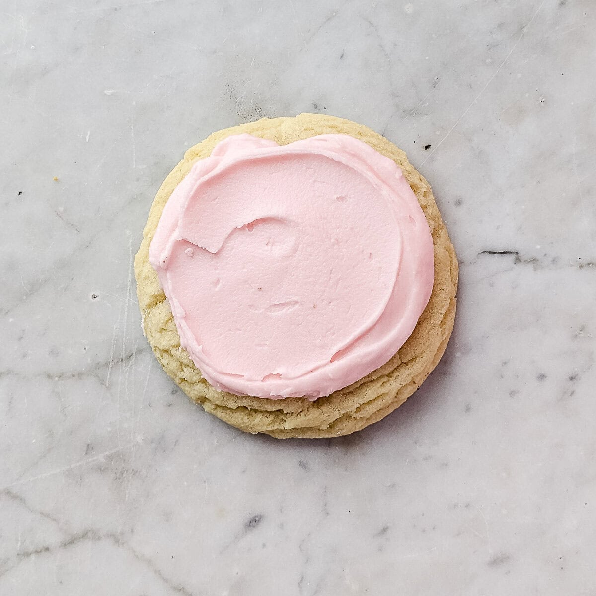A Crumbl sugar cookie frosted with light pink frosting on a marble surface background.