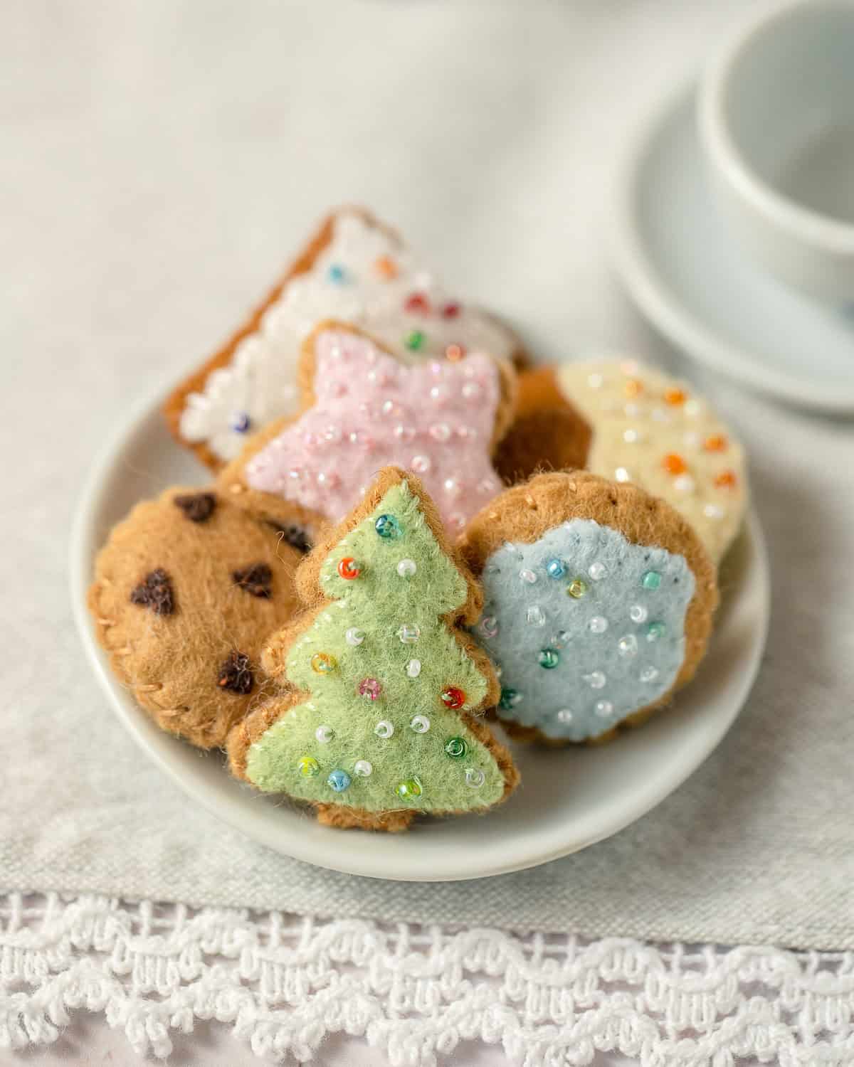 A mini plate filled with six tiny felt cookies: a Christmas tree, a chocolate chip cookie, a dipped cookie, a round frosted cookie, a rectangle frosted cookie, and a star-shaped frosted cookie.
