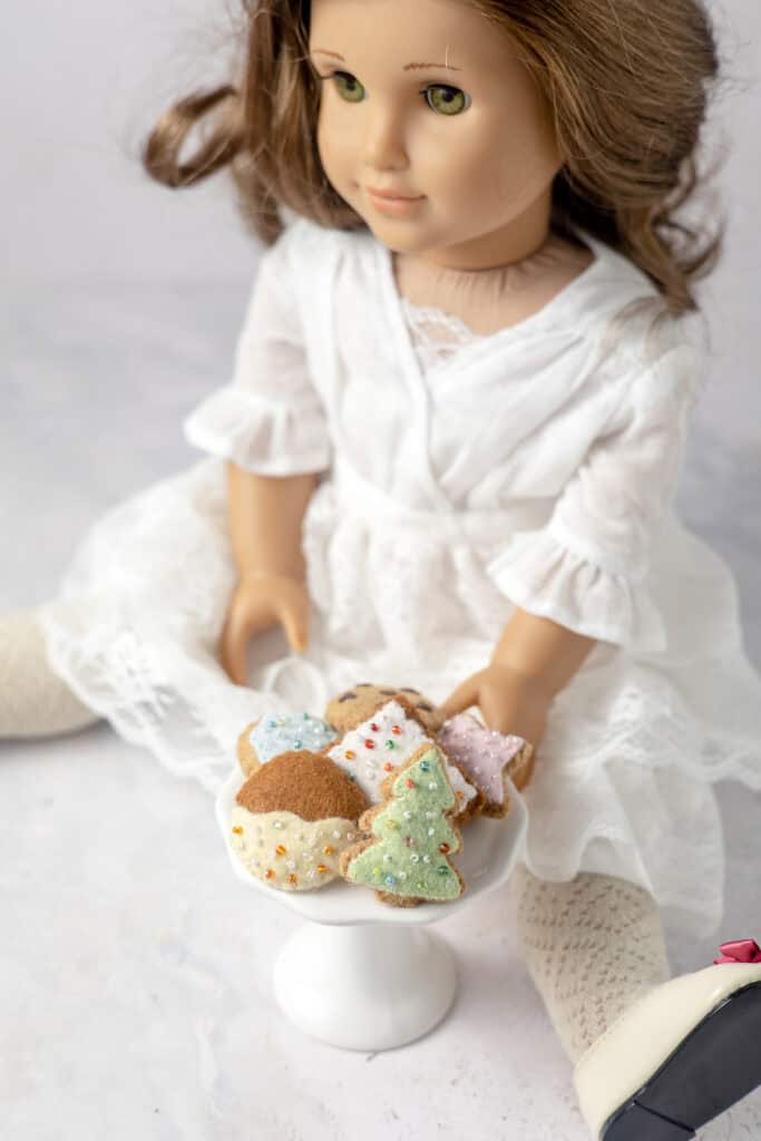 An 18" doll sitting beside a tiny cake stand filled with mini felt cookies including a dipped cookie, a Christmas tree, and a white-frosted rectangle cookie.