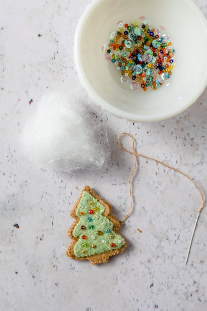 Stitching a green tree-shaped piece of felt decorated with beads onto brown pieces of tree-shaped felt with a piece of poly stuffing and dish of seed beads nearby.