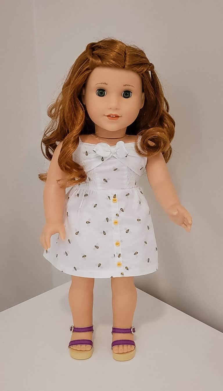 An American Girl doll with red hair, white dress with bumblebees and purple sandals.