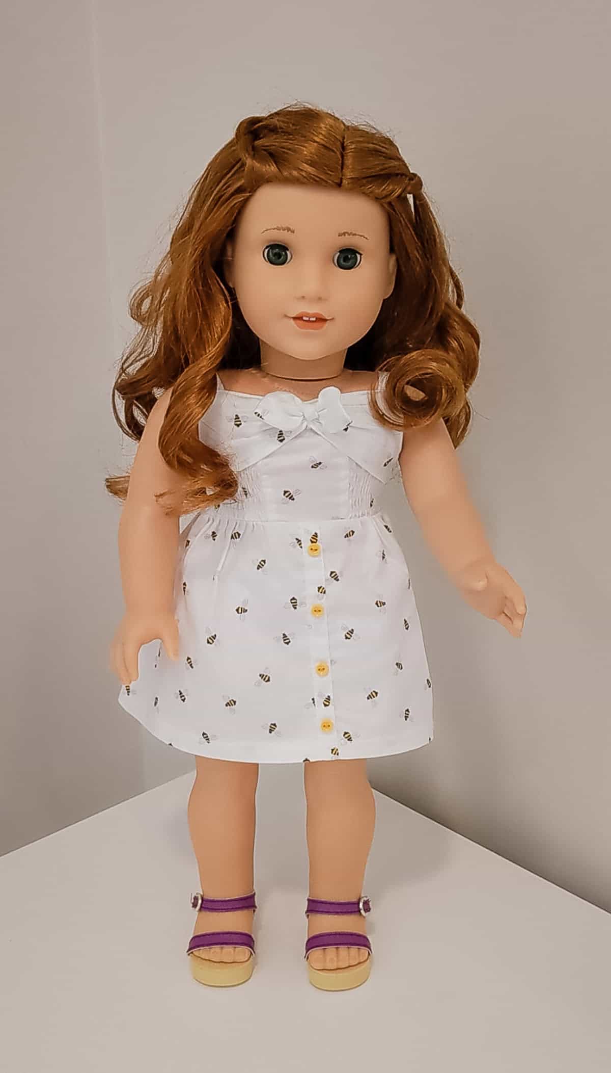An American Girl doll with red hair, white dress with bumblebees and purple sandals.